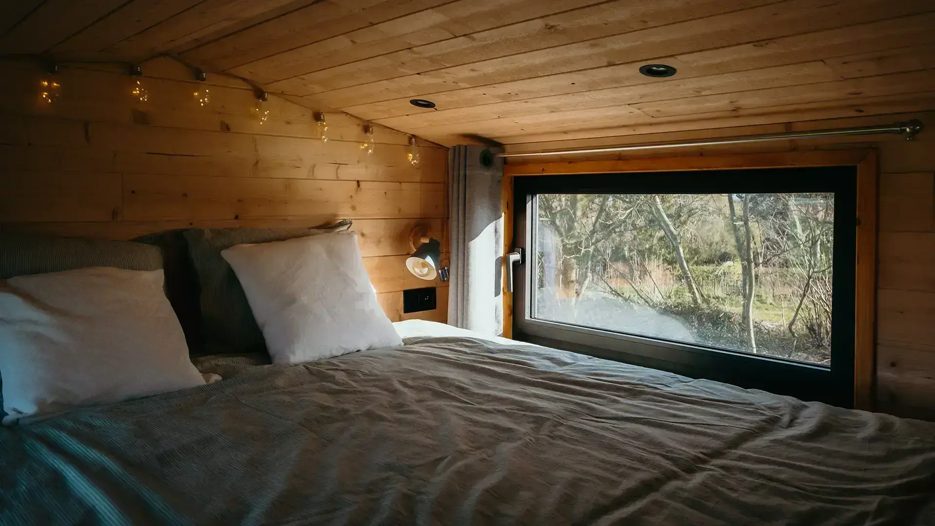 Lit double avec un drap de lit Kaki face à une petite fenêtre dans une tiny house