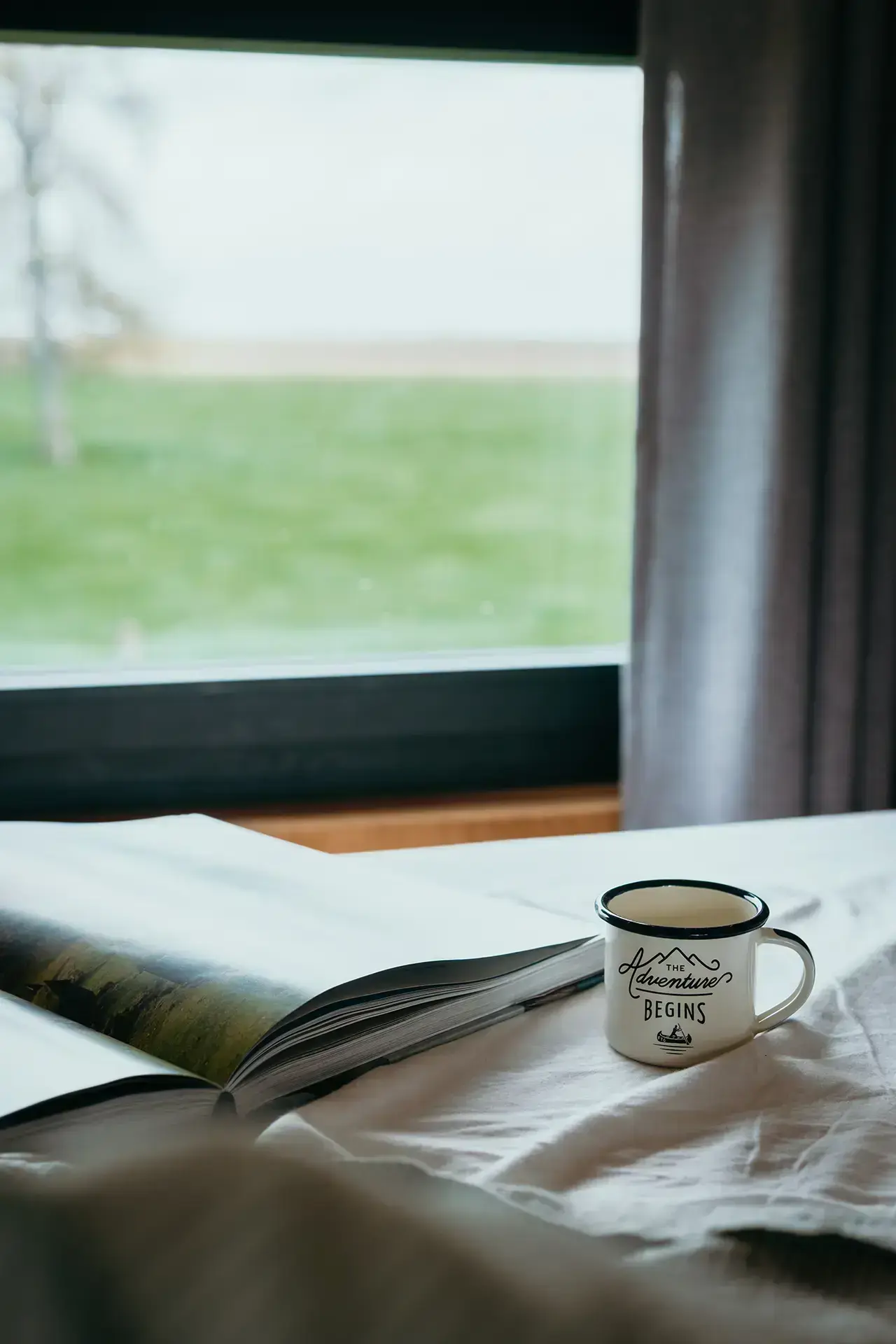 Tasse de café et livre d'aventure ouvert avec la vue sur l'extérieur depuis le lit d'une Tiny House